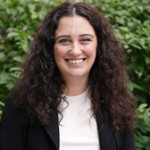 A woman with curly dark hair smiles in front of a bush wearing a white shirt and black jacket.