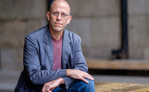A man wearing glasses in jeans and a blazer poses while leaning with his hands on his knee.