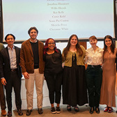A row of six well-dressed students pose in front of a projector screen with their names on it.